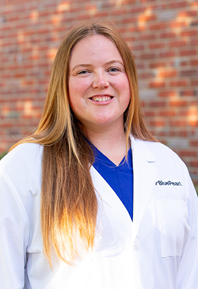 Dr. Julia Matsen smiles outside in front of a brick building. She is wearing blue scrubs and her BluePearl whitecoat.