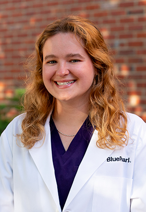 Dr. Chloe Roberts smiles outside in front of a brick building. She is wearing purple scrubs and her BluePearl whitecoat.