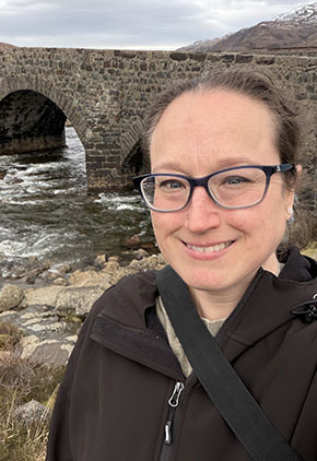 Jess Shissler smiles outside in Ireland. A bridge over a flowing river is behind her. She is wearing a black jacket with a black crossbody bag.
