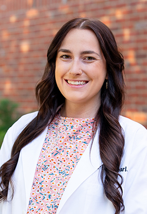 Dr. Taylor Ayars smiles outside in front of a brick building. She is wearing her BluePearl whitecoat and a light pink blouse that features small blue and white flowers.
