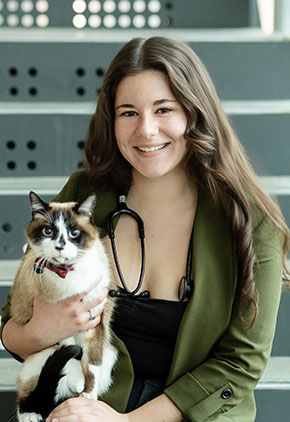 Dr. Ariel Ackerman smiles while sitting outside on metal steps. She has a stethoscope around her neck and is wearing a black top and olive green blazer while holding her brown and white cat.