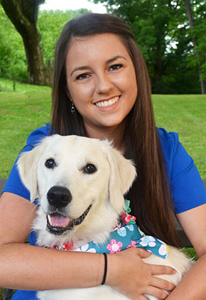 Dr. Ashton Haas smiles outside in a expansive green space. She is sitting with her arms around her golden retriever in her lap. She has on a blue t-shirt.