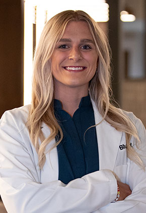Dr. Kylie Levan smiles for a headshot in the lobby of BluePearl Pet Hospital. She is wearing a black blouse and her whitecoat with her arms crossed across her body.
