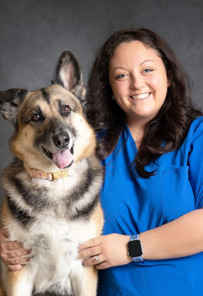 Dr. Jonna Woodson smiles for a professional photo with her German shepherd. They are posed side-by-side and Dr. Woodson is wearing blue scrubs.