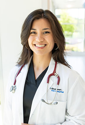 Dr. Ashley Broderick smiles in the windowed lobby of BluePearl Pet Hospital. She is wearing black scrubs, her whitecoat and has a stethoscope around her neck.