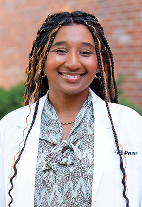 Dr. Ariana Cook smiles outside in front of a brick building. She is wearing a light blue patterned blouse and her BluePearl whitecoat.