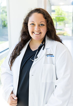 Dr. Jonna Woodson smiles in the windowed lobby of BluePearl Pet Hospital. She is wearing black scrubs, her whitecoat and has a stethoscope around her neck.