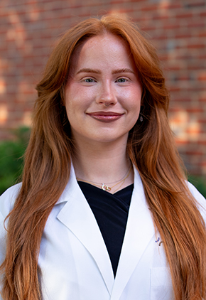 Dr. Kaley Boone smiles outside in front of a brick building. She is wearing black scrubs and her BluePearl whitecoat.