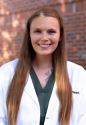 Dr. Justine Finerty smiles outside in front of a brick building. She is wearing a forest green blouse and her BluePearl whitecoat.
