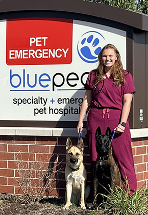 Dr. Justine Finerty smiles outside next to the exterior BluePearl Pet Hospital sign at our Oak Creek hospital. She is holding the leashes of her two dogs and wearing red scrubs.