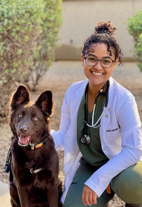 Dr. Sirah Javier smiles outside while kneeling next to her black dog. She is wearing olive green scrubs, her whitecoat and she has a stethoscope around her neck.