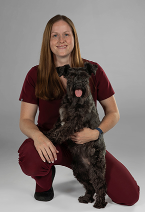 Dr. Lindsey Schmeiser smiles for a professional studio headshot. She is wearing maroon scrubs and kneeling beside her dog.