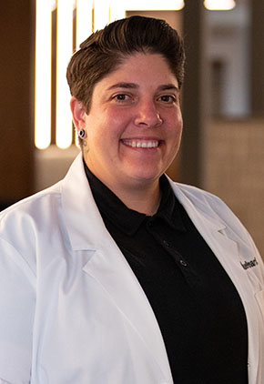 Dr. Rania Abouzedan smiles for a headshot in the lobby of BluePearl Pet Hospital. She is wearing a black top and her whitecoat.