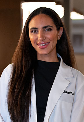 Dr. Stephanie Malouf smiles for a headshot in the lobby of BluePearl. She is wearing a black top and her whitecoat.