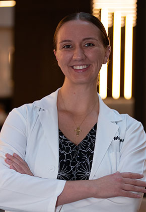Dr. Christine Cassen smiles for a headshot in the lobby of BluePearl Pet Hospital. She is wearing her whitecoat and a black blouse that features white outlines of flowers.