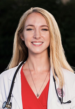 Dr. Grace Johnston smiles outside for a headshot. She is wearing her St. George's University whitecoat, a coral blouse and has a stethoscope around her neck.