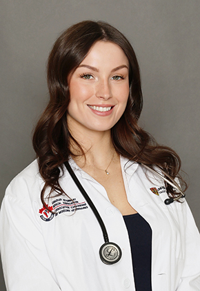 Dr. Rebecca Wassmuth smiles for a professional headshot in front of a grey background. She is wearing her university whitecoat and has a stethoscope around her neck.