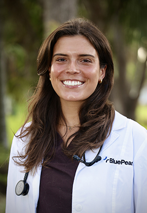 Dr. Carolina Graham smiles outside for a professional headshot. She is wearing dark brown scrubs, her BluePearl whitecoat and has a stethoscope around her neck.
