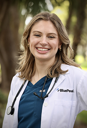 Dr. Samantha Higgins smiles outside for a professional headshot. She is wearing blue scrubs and her BluePearl whitecoat with a stethoscope around her neck.