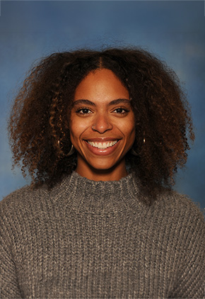 Dr. NaaLamiley Kwakye smiles for a professional headshot in front of a blue backdrop. She is wearing a grey sweater.