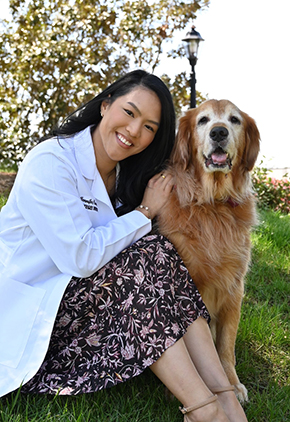 Dr. Jennifer Maharath smiles outside sitting on a small hill with her golden retriever. She is wearing a floral dress and her Tuskegee whitecoat.