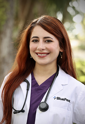 Dr. Amanda Rosendo Ocasio smiles outside for a professional headshot. She is wearing purple scrubs, her BluePearl whitecoat and has a stethoscope around her neck.