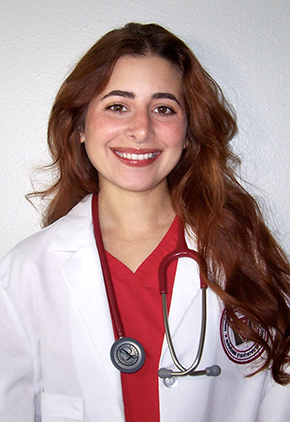 Dr. Amanda Rosendo Ocasio smiles for a headshot in front of a white background. She is wearing her whitecoat and red scrubs with a stethoscope around her neck.