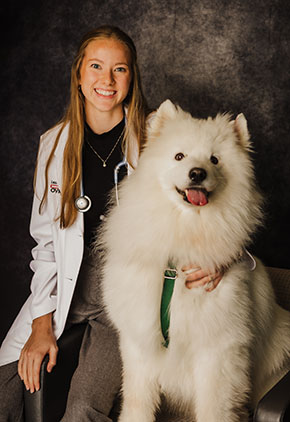 Dr. Lexi Wheeler smiles while sitting next to her Samoyed dog for a studio portrait. She is wearing dark grey pants a black top and her whitecoat with a stethoscope around her neck.