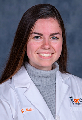 Dr. Grace Malla smiles for a professional headshot in front of a blue-grey background. She is wearing a grey turtleneck and her UT white coat.