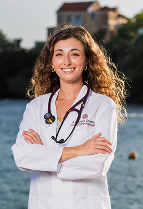 Dr. Gabriella Bonofiglio smiles outside on the beach in Grenada. She is wearing her St. George's University whitecoat and has a stethoscope around her neck.