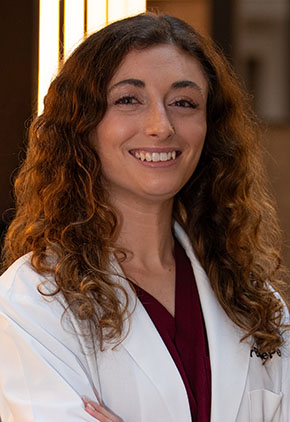Dr. Gabriella Bonofiglio smiles for a headshot in the lobby of BluePearl Pet Hospital. She is wearing maroon scrubs and her whitecoat.