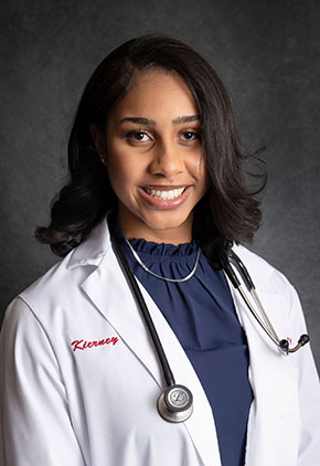 Dr. Kierney Burks smiles for a studio headshot in front of a grey backdrop. She is wearing a navy blouse and her whitecoat. She has a stethoscope around her neck.