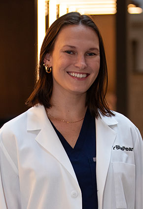 Dr. Brittany Whitcher smiles for a headshot in the lobby of BluePearl Pet Hospital. She is wearing black scrubs and her whitecoat.