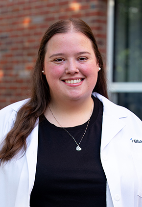 Dr. Madison Reed smiles outside in front of a brick building. She is wearing a black blouse and her BluePearl whitecoat.