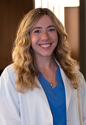 Dr. Kyra Boss smiles for a headshot in the lobby of BluePearl Pet Hospital. She is wearing blue scrubs and her whitecoat.