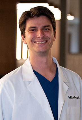 Dr. Grant Lorimer smiles for a headshot in the lobby of BluePearl. He is wearing navy scrubs and his whitecoat.