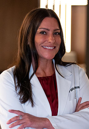 Dr. Mirelle Syrja smiles for a headshot in the lobby of BluePearl Pet Hospital. She is wearing a red blouse and her whitecoat.