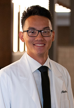 Dr. Erik Yang smiles for a headshot in the lobby of BluePearl Pet Hospital. He is wearing a white dress shirt, black tie and his whitecoat.