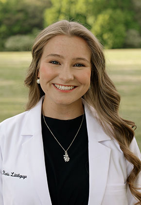 Dr. Karis Littlepage smiles outside in an expansive green space, She is wearing a black top and her whitecoat.
