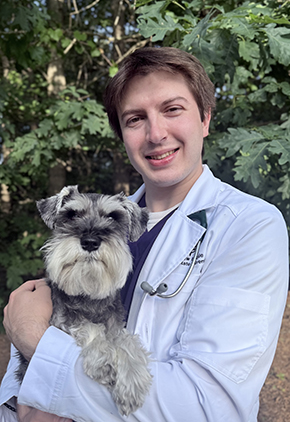 Dr. Matthew Mackie smiles outside in front of a leafy tree. He his holding his grey and white miniature schnauzer in his arms and is wearing whitecoat.