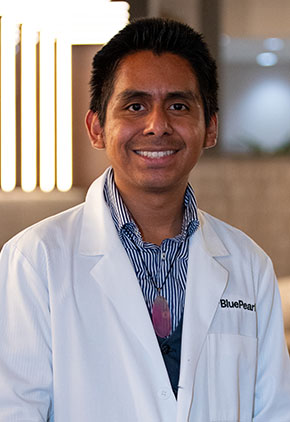 Dr. Sean Garcia smiles for a headshot in the lobby of BluePearl. He is wearing a blue button-up and his whitecoat.