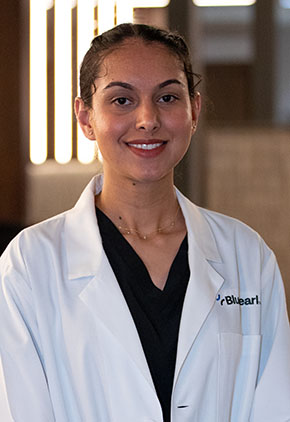 Dr. Muskanjot Makkar smiles for a headshot in the lobby of BluePearl Pet Hospital. She is wearing black scrubs and her whitecoat.