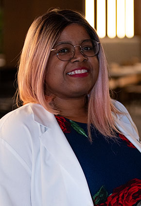 Dr. Kandice Mills smiles for a headshot in the lobby of BluePearl Pet Hospital. She is wearing a navy blouse that features red roses and her whitecoat.