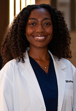 Dr. Imara Savon smiles for a headshot in the lobby of BluePearl Pet Hospital. She is wearing navy scrubs and her whitecoat.