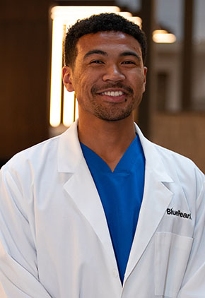 Dr. Jason Simmons smiles for a headshot in the lobby of BluePearl Pet Hospital. He is wearing blue scrubs and his whitecoat.