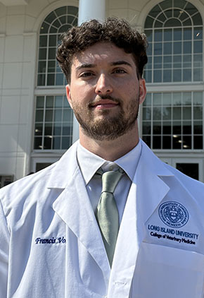 Dr. Francis Volz IV smiles outside Long Island University School of Veterinary Medicine. He is wearing a white dress shirt, light silver-green tie and his school whitecoat.