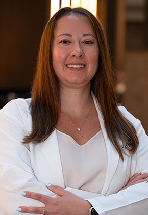 Dr. Allison Klenk smiles for a headshot in the lobby of BluePearl Pet Hospital. She is wearing a taupe blouse and her whitecoat with her arms crossed across her body.