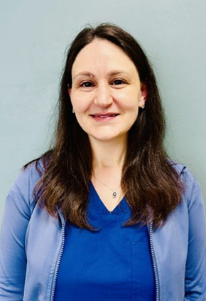 Dr. Ellyn Gouldin smiles for a headshot in front of a grey background. She is wearing royal blue scrubs and a powder blue cardigan.