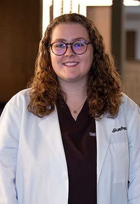 Dr. Kylie McClure smiles for a headshot in the lobby of BluePearl Pet Hospital. She is wearing maroon scrubs and her whitecoat.