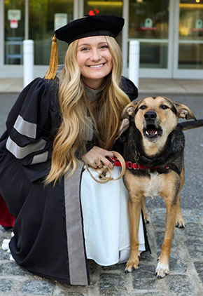 Dr. Haley Zeliff smiles outside while squatting next to her black and tan mixed-breed dog. She is in her doctoral robes.